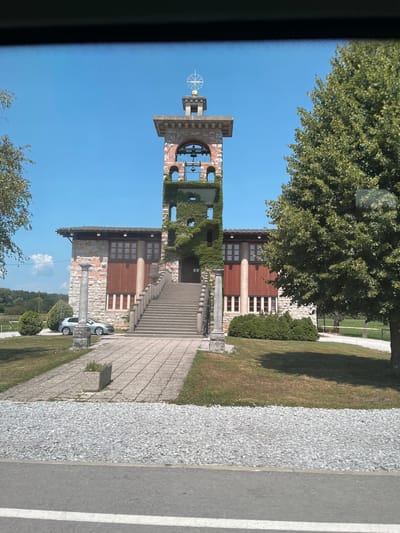 The image shows the Church of the Holy Spirit in Črna vas, Slovenia. It is a bright, sunny day with a clear blue sky. The church has a distinct bell tower covered in ivy, with a golden dome and a cross at the very top. In front of the bell tower is a large set of stairs leading up to the entrance of the church. The church building itself is made of stone and wood, with large windows. To the left of the church, a silver car is parked on a paved pathway. The area around the church is grassy, with some bushes and trees. The foreground shows a gravel path and a road with a white line.