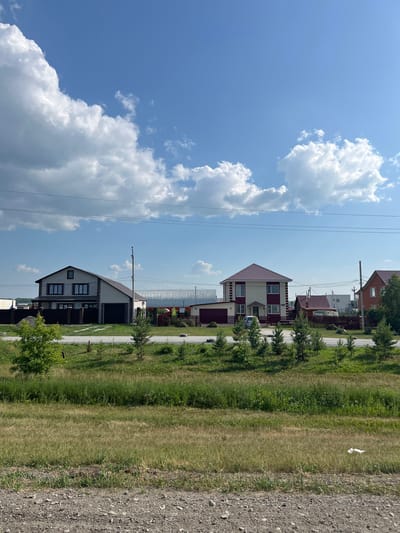 The image depicts a suburban street scene in Prokudskoye, Russia, on a clear, sunny day. The foreground is dominated by a gravel road and a strip of dry, sparse grass. Beyond this, a line of young evergreen trees runs parallel to a paved road.
Along the paved road, several residential houses are visible, separated by wooden fences. The houses are of various designs and colors, with pitched roofs. One house on the left is light-colored with dark trim and a dark garage. To its right, a larger, two-story house features a light-colored facade with maroon accents and a maroon roof. Further to the right, another house has a brick facade and a red roof. In the background, larger industrial-looking structures and more houses are present.
The sky is a bright blue with scattered white cumulus clouds. Power lines stretch across the upper portion of the image. The overall impression is one of a developing residential area on the outskirts of a town, characterized by new construction and open spaces.