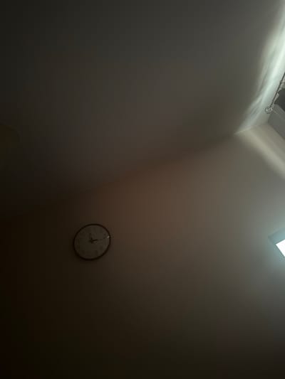 The image shows a close-up view of a round analog clock mounted on a plain, light-colored wall. The clock has a dark bezel and a white face with black numbers and hands. The time displayed on the clock is approximately 10:42. The lighting in the image is dim, with a bright shaft of light entering from the upper right corner, casting shadows and illuminating the upper portion of the wall and ceiling. The scene is indoors, likely a room, with no other objects or people clearly visible. The location is specified as Tuszynek Majoracki, Poland.