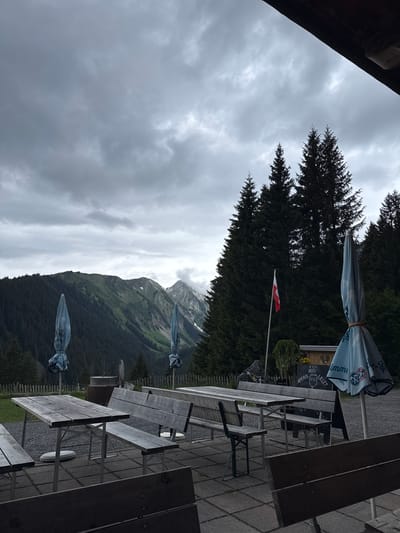 The image depicts an outdoor patio area in a mountainous setting, likely in Untergiblen, Austria, as indicated by the context. The scene is dominated by several wooden picnic-style tables and benches arranged on a paved surface. Two large, closed outdoor umbrellas, one displaying a logo that appears to be "SCHWEPPES" or "Unicum," are present, suggesting a restaurant or cafe patio.

In the midground, a red and white flag, identifiable as the Austrian flag, flies from a flagpole. A menu board or sign is partially visible near one of the umbrellas, displaying text such as "LILLET," "HUND," "IMPERIAL," and prices like "€ 7.50" and "0.50."

The background features dense evergreen forests covering the lower slopes of rugged mountains. The mountain peaks, some with visible snow patches, are partially obscured by thick, grey overcast clouds, indicating cloudy weather. No people are present, and the closed umbrellas suggest that the patio is not currently in use, possibly due to the weather or time of day. The overall atmosphere is quiet and subdued.