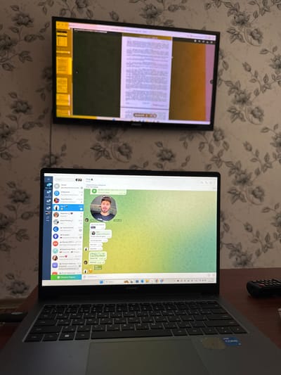 The scene depicts an indoor room in Ciorescu, Moldova. A dark brown desk occupies the foreground, upon which a dark gray Huawei laptop is open. The laptop's screen displays the Telegram desktop application, showing a list of chats and contacts in Cyrillic on the left panel. The main chat area features a profile picture of a young male with a beard and dark hair, smiling, alongside multiple messages written in Cyrillic against a green-yellow gradient background. The laptop's keyboard features a QWERTY layout with Cyrillic characters, and an "Intel Core i7" sticker is visible on the palm rest. To the right of the laptop, a black remote control is partially visible on the desk surface.
The wall behind the desk is covered with light-colored wallpaper featuring a repeating pattern of dark gray floral designs. A black flat-screen television is mounted on this wall, positioned above the laptop. The television screen displays what appears to be a document or webpage, with text prominently featured on the right side against a light background, and a darker, yellowish panel on the left. A control interface or progress bar is visible at the bottom center of the TV screen. A dark cable runs vertically down the wall from behind the television. No physical individuals are present in the immediate scene.