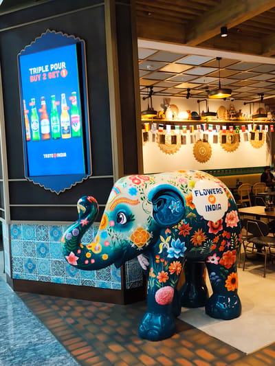 The image captures a vibrant scene inside a establishment in Al Matar, United Arab Emirates. In the foreground, a large, artistically painted fiberglass elephant statue is the central subject. The elephant is adorned with a colorful array of floral patterns and intricate designs in shades of blue, teal, orange, pink, and yellow. The text "FLOWERS OF INDIA" is prominently displayed on its side.
Behind the elephant, a bright blue digital billboard displays a promotional advertisement that reads "TRIPLE POUR BUY 2 GET 1". Below this text, images of several beer bottles are visible. The billboard is framed by a decorative blue border with a scalloped edge.
The environment suggests an indoor dining or retail space. The background features a ceiling with a grid of wooden panels and lighting fixtures. Further back, a row of small international flags is strung across the wall. Restaurant seating with tables and chairs is visible, and a few patrons are seated at tables, with one individual in the background appearing to be an employee. The flooring consists of patterned tiles and a darker brick-like material. The overall atmosphere is lively and visually engaging, likely a restaurant or bar with a thematic decor.