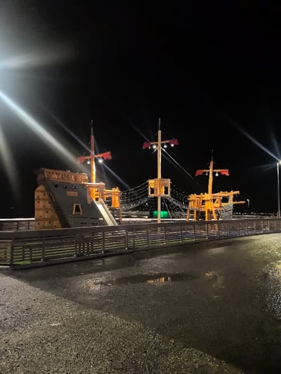 The image displays a pirate ship-themed playground located in Sjøvegan, Norway, at night. The foreground consists of a dark, wet paved surface with several visible puddles reflecting light. A horizontal wooden slatted fence, dark in color, runs across the midground, separating the paved area from the playground structure. The primary subject is a large, multi-level playground constructed from wood and gray-painted elements, designed to resemble a pirate ship. It features three prominent wooden masts, each topped with a red, banner-like structure and equipped with artificial floodlights. The playground incorporates slides, a climbing wall on its left side, and multiple interconnected rope net bridges. A distinct green light source is visible beneath one of the rope net sections. Illumination is provided by the playground's internal lights and two external streetlights; one on the far left casts a prominent lens flare, and another on the far right contributes to the overall scene lighting. The background is a uniformly dark night sky. No individuals are present within the frame.