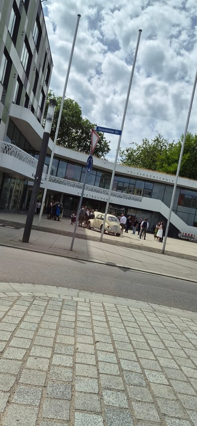 The image captures an outdoor scene at Rosenheimer Str. 30 in Kolbermoor, Germany, featuring the Volkshochschule and Stadtbücherei building on a cloudy day.
In the foreground, grey square cobblestones form a paved area, transitioning to an asphalt road and then a light-colored paved sidewalk leading to the buildings. A dark lamppost stands prominently on the left side of the sidewalk. Three tall, slender, silver-colored flagpoles are erected along the sidewalk, extending towards the sky.
On the main building, the text "STADTBÜCHEREI" and "VOLKSHOCHSCHULE" is visible in white letters, identifying the institutions. The building itself is modern, with large glass windows on the lower level, a white exterior, and a distinctive curved architectural element above the ground floor, featuring decorative cut-outs. A green roof with visible vegetation covers the building. To the left, a portion of an adjacent, multi-story building with a grey and light-colored paneled facade and rectangular windows is visible.
A street sign with "Rosenheimer Str." in white text on a blue background is affixed to one of the flagpoles, accompanied by a white triangular yield sign with a red border and a blue circular mandatory sign with a white arrow.
Numerous individuals are gathered on the sidewalk in front of the Volkshochschule/Stadtbücherei. Several people are dressed in traditional Bavarian attire, including women in Dirndls and men in Lederhosen, suggesting a cultural event or celebration. Other individuals wear formal or semi-formal clothing, such as suits and dresses. A light-colored vintage Volkswagen Beetle is parked on the sidewalk, with its front hood open. The vehicle's license plate reads "AUB XS938". A child is visible standing near the car.
The sky above is partially overcast with white and grey clouds. Green trees are visible behind and on the roof of the main building. A red and white striped barrier or container is partially visible on the far right side of the scene.