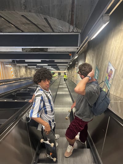 Two young men are on an escalator in a subway station. The man on the left has dark, curly hair and is wearing a white and blue striped shirt and black shorts. He is holding a skateboard in his right hand. The man on the right has light brown, curly hair and is wearing a grey t-shirt, maroon shorts, and white sneakers. He has a blue backpack on and is holding his right hand to the back of his head. The escalator is going down, and the walls of the station are made of concrete and rock. There are posters on the wall to the right. The station appears to be clean and well-lit. The overall impression is of a modern and functional public transportation hub. The location is Råcksta metro station in Solna, Sweden.