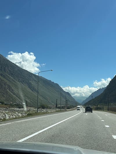 This is a horizontal, eye-level, long shot of a highway in the city of Upper Baksan, Russia. The highway is paved and has white lane markings. There are two cars driving on the highway, one white van in the distance and a black SUV in the foreground. The highway is surrounded by mountains on both sides. The mountains are covered in greenery and there are some clouds in the sky. The sky is blue and there are some white clouds. The scene is likely captured during the daytime.