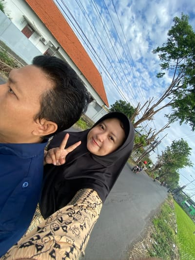 The image displays an outdoor scene on a paved road in Putat, Indonesia. In the immediate foreground, two individuals are present. A male, visible from the left side of his head and torso, wears a blue collared shirt and faces left, away from the viewer. To his right, a female wearing a black hijab and a light-colored, patterned long-sleeved garment smiles directly at the camera, extending her right hand to form a peace sign.

The background features a building with a white facade and an orange-tiled roof on the left. Numerous overhead utility lines crisscross the blue sky, which contains scattered white clouds. The asphalt road extends into the distance, bordered by trees, some with bare branches, on the right. Farther down the road, at least two motorcycles are visible, one with a rider wearing a red helmet. Green vegetation, consistent with agricultural fields, is present along the right edge of the distant road.