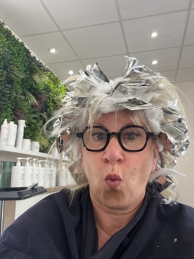 A selfie taken inside a hair salon in Montrond-les-Bains, France shows a woman with foil and cotton in her hair as part of a hair coloring process. She is wearing black-rimmed glasses and has her lips pursed. The background includes a wall with artificial greenery, bottles of hair care products, and a white tiled ceiling with recessed lights.