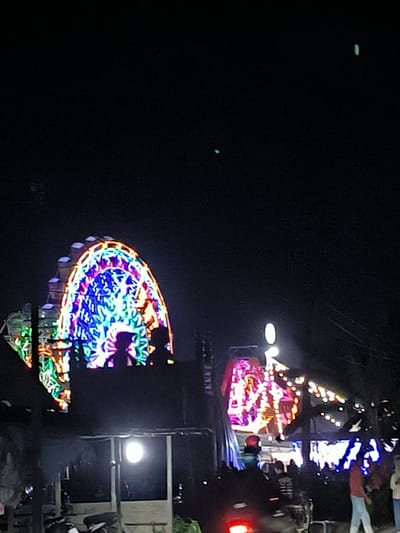 The image shows an outdoor scene at night in Blangbitra, Indonesia, where carnival rides, including a large Ferris wheel, are illuminated with vibrant lights. The Ferris wheel, prominently displayed on the left, features a colorful display of lights in various hues. In the foreground, several people are visible, some of whom appear to be standing or walking amidst a collection of parked motorcycles and structures. A separate, smaller structure is lit up with lights in the center of the shot, with additional strings of lights extending behind it. The setting suggests a fair or carnival atmosphere.