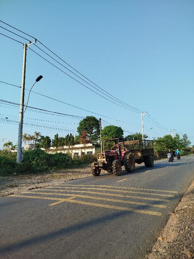 The image depicts a rural or semi-rural road scene in Đăk Xú, Vietnam, under a clear blue sky. A red, open-cabin tractor with a loaded, wooden-sided trailer is traveling from left to right on the asphalt road, positioned near the center-left. The trailer appears to contain dark material or soil. Two motorcycles are visible behind the tractor, traveling in the same direction on the right side of the road. The lead motorcycle is white, ridden by a single individual, followed by a darker motorcycle also ridden by one person.
The road features several sets of parallel yellow lines, possibly indicating speed bumps or rumble strips, extending across the lane where the tractor is located. Shadows are cast across the road surface. To the left of the road, a concrete utility pole with a street lamp and multiple overhead power lines stands prominently. Dense green foliage and trees line both sides of the road, with a white building partially visible behind the vegetation in the mid-ground on the left. Another utility pole with lines is visible further down the road on the right. The overall lighting suggests a bright, sunny day.
