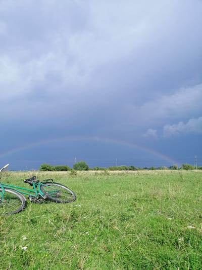 The image depicts an outdoor scene with a green bicycle lying on its side in a grassy field. The bicycle is positioned in the foreground, angled diagonally from the lower left to the upper right of the frame. Behind the bicycle, the field continues, dotted with patches of dry grass and small white flowers. In the middle ground, a line of trees and shrubs forms a distant horizon. Above the trees, the sky is dramatic, with dark, stormy clouds on the left side of the image and lighter clouds on the right. A rainbow arcs across the sky, visible above the tree line, with its colors muted against the dark clouds. A single utility pole with wires is visible on the right side of the image. The overall impression is a natural landscape under a transitioning weather system, with the bicycle suggesting a moment of rest or an abandoned journey. The location is identified as Krasnoluka, Ukraine.