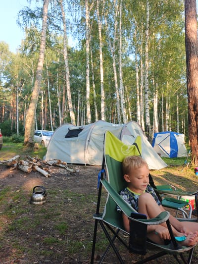 The image depicts a campsite in Nepryakhinskoye Sel'skoye Poseleniye, Russia. A young boy is seated in a green camping chair, holding a blue plastic fork. In the background, several tents are pitched amongst birch and pine trees. A stack of firewood lies on the ground near a silver kettle. A white car is parked behind one of the tents. The scene is illuminated by natural sunlight filtering through the trees, creating dappled shadows on the ground.
