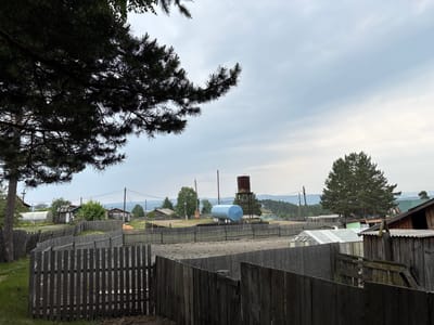 The image depicts a rural scene in the village of Nevon, Russia. In the foreground, a weathered wooden fence runs horizontally across the frame, with another fence line extending further into the distance. Behind the fences, a dusty, open area is visible. A large, light blue cylindrical tank is mounted on a metal structure in the middle ground, accompanied by a smaller, rusty water tower. Several wooden buildings, some with visible greenhouses, are scattered throughout the landscape. Power lines and utility poles are present. In the background, rolling hills and a treeline can be seen under an overcast sky. A large pine tree branch with needles and cones overhangs the top left portion of the image. The overall impression is one of a quiet, somewhat aged rural settlement.