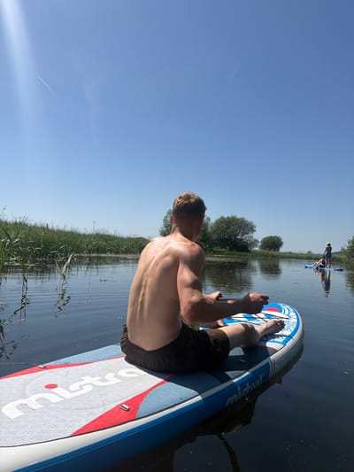 A man is seated on a paddleboard on a body of water, possibly the River Stour, as Babergh is near this river in the United Kingdom. The paddleboard is blue and white, with the word "Mistral" visible on its surface. The man has fair skin and short blonde hair. He is shirtless, wearing dark-colored shorts, and holding a paddle. Another paddleboarder is visible in the distance. The background consists of trees and reeds along the shoreline. The sky is a clear blue.