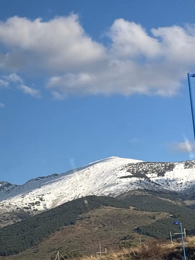 The image displays a panoramic landscape of the snow-capped Sierra de Guadarrama mountain range, observed from the vicinity of Otero de Herreros, Spain. The upper half of the frame is dominated by a clear, bright blue sky with several scattered white cumulus clouds. Below, the highest visible mountain peaks are covered in extensive white snow. Mid-slopes reveal a transition from snow to dark green conifer forests, which form distinct bands across the terrain. The lower mountain slopes and immediate foreground consist of brown, dry-looking scrubland with sparse vegetation and visible rocky patches.

In the foreground, on the right side, a tall, opaque blue vertical structure, possibly a pole or part of a larger amenity, is partially visible. Further down and to the right, a smaller blue streetlamp-like fixture on a thin pole is present. Indistinct metallic structures or fence posts are also discernible in the brown foreground terrain. No individuals or animate subjects are depicted. The scene is brightly illuminated, indicating daylight conditions. A faint reflection, likely from a window, is present in the lower center of the image.