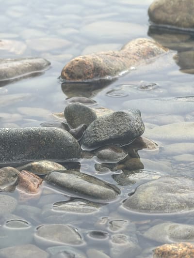 The image depicts a close-up view of numerous smooth, water-worn stones submerged in clear, shallow water. The stones vary in size, shape, and color, ranging from light grays and beiges to darker, mottled browns and grays. Some stones are partially covered by a thin layer of water, creating reflections and highlights. The water appears calm, with subtle ripples on the surface. The scene is natural and appears to be an outdoor environment, likely a riverbed or shoreline. There are no people, animals, or man-made objects visible in the image. The location in Ust-Kabirza, Russia, suggests a natural setting such as a river or lake within or near the city.