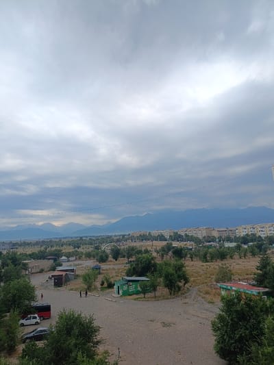 This image depicts an overcast day in Tokmok City, Kyrgyzstan, with a view of mountains in the background. In the foreground, there is a gravel parking area with several vehicles including a white car, a dark sedan, and a red bus. A few small buildings, possibly storage units or kiosks, are visible in the middle ground. There are also scattered trees and vegetation throughout the scene. Several people are present in the parking area, with some individuals walking and others standing. In the distance, there is a row of apartment buildings and more trees, with the snow-capped mountains forming a dramatic backdrop. The sky is filled with heavy, gray clouds, suggesting a possibility of rain.