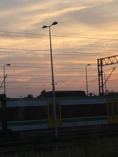 The image depicts a train station in Burkat, Poland, at dusk. A train with white and green livery is visible, with yellow doors and the number "2" displayed. Overhead, train power lines stretch across the frame, supported by poles and an industrial metal structure. Several streetlights stand along the platform, silhouetted against a sky filled with soft, pastel clouds in shades of orange and pink. The foreground is dominated by the train tracks and a strip of tall, dark grass. No people are visible in the scene.