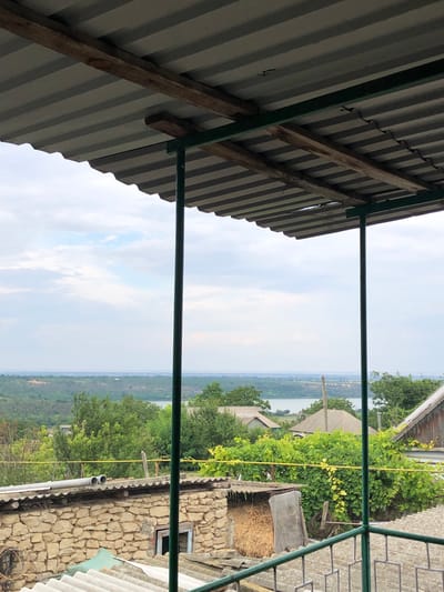 This is a medium shot, captured from a covered patio, looking out at a rural landscape under an overcast sky. In the foreground, the corrugated metal roof of the patio is visible, supported by green metal poles and wooden beams. Below the patio, a rustic stone building with a hay-filled alcove is apparent. Beyond the immediate structures, a lush green expanse of trees and vegetation leads to a body of water in the distance, which appears to be a river or lake. Several houses with pitched roofs are scattered throughout the landscape. The scene is set in the village of Calfa, Moldova, suggesting a residential or agricultural setting. There are no people visible in the image.