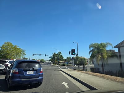 The image shows a view from inside a vehicle driving on a sunny day. The scene is a suburban street in West Sacramento, California, with a clear blue sky and some scattered clouds. The foreground features the rear of a blue Honda Fit. In the middle ground, there are traffic lights, some of which are green, indicating traffic is moving. A street sign indicates "Evergreen Ave". The background shows buildings, palm trees, and other vehicles, suggesting a commercial or residential area. The time of day appears to be late morning or early afternoon, given the bright sunlight and long shadows. There is a white arrow painted on the road, indicating a straight direction of travel. The license plate on the Honda Fit reads "7MTG362 California". The overall impression is a typical day in a suburban environment with traffic and clear weather.