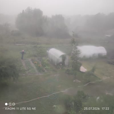 The image depicts a foggy, outdoor scene in the village of Bayunovskie Klyuchi, Russia. In the foreground, a scarecrow stands in a grassy area with a path leading away from it. To the right of the scarecrow, there are some yellow flowers. Further into the scene, there are two greenhouses. One is a translucent, arched structure in the middle ground, and another is a larger, similarly shaped greenhouse partially obscured by fog in the background. The ground appears to be damp, with what looks like a garden hose and sprinklers visible, suggesting recent watering. The overall atmosphere is hazy and overcast. Text at the bottom of the image indicates the camera used was a "XIAOMI 11 LITE 5G NE" and the date and time are "23.07.2026 17:22".