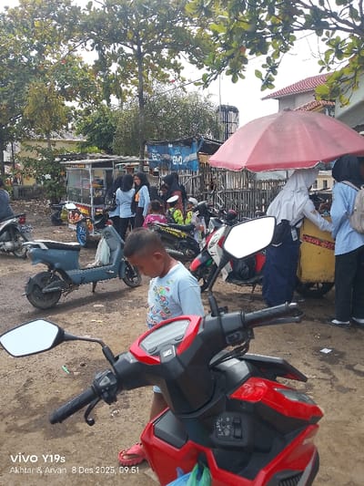 A bustling outdoor scene shows a young boy standing near a red Honda motorcycle, its mirror prominently displayed. Nearby, schoolgirls in blue shirts and hijabs gather near a food vendor's cart sheltered by a red umbrella. Various motorcycles and an electric scooter are parked around, adding to the lively atmosphere. Other children and adults mingle in the background, suggesting a busy local market or school setting.