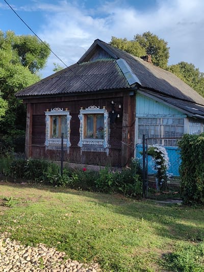 The image depicts a rustic, wooden house with a corrugated metal roof, situated in a rural setting, likely a dacha or village home in Квант-88 Снт, Russia. The house is weathered, with dark brown wooden walls. It features two intricately carved white window frames on the left side, each with a yellow and blue decorative trim, and two panes of glass. The right side of the house has a light blue facade and a large, multi-paned window. Green foliage, including dense bushes and trees, surrounds the house. A wire fence runs along the front of the property, partially obscured by plants. In the foreground, there is a patch of green grass and a strip of light-colored gravel. A single red rose is visible in the vegetation near the fence. The sky above is blue with scattered white clouds. There are no people visible in the image.