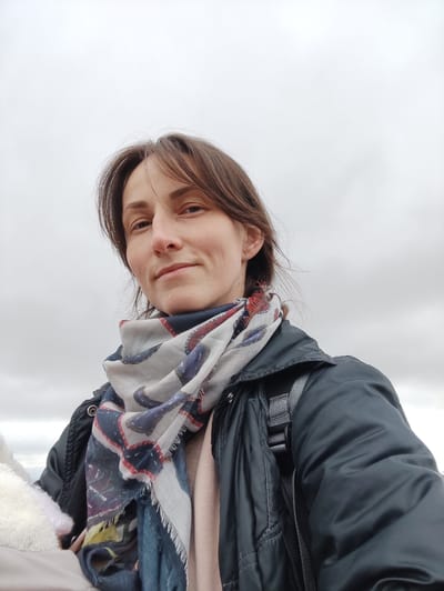 The image depicts a close-up, low-angle portrait of an adult female outdoors in Bestuzhevskoye, Russia. The subject, positioned centrally, has light skin, brown hair, and appears to be in her 30s or 40s. She is looking directly at the camera with a neutral expression, exhibiting a slight smile. Her left ear displays a small, silver hoop earring. She is dressed in a dark grey or black quilted jacket worn over a light pink or beige layer. A multi-colored scarf with patterns in blue, red, and white is wrapped around her neck. A grey strap from a backpack is visible over her right shoulder. To the lower left, a portion of a white, textured, possibly fuzzy object is visible. The background consists entirely of a light grey, overcast sky.