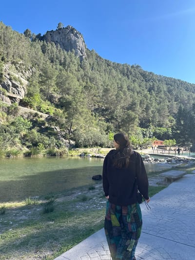 A full shot, slightly rotated to the left, shows a woman standing near a river and a cliff in Montanejos, Spain.
The woman, positioned in the right of the shot, is wearing a black sweater and a patterned skirt. She is facing away from the cliff and appears to be looking down at the river.
The river runs vertically through the center of the image, with green grass and foliage on its banks. A gray stone pathway is on the right.
On the left, a cliff covered in trees rises up to the sky. The sky is a clear blue. In the background, a bridge-like structure can be seen.