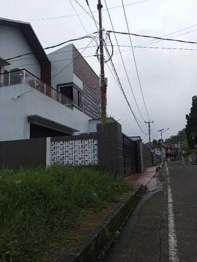 A modern, multi-level house with white and brown facades stands behind a dark gray fence with a patterned white section. Lush green grass grows wildly along the curb. A utility pole with numerous wires dominates the view. A narrow paved street with a white line marker stretches into the distance, lined with houses and greenery under an overcast sky.
