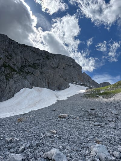 The image depicts a mountainous landscape with a large, steep rock face on the left, partially covered with patches of green vegetation. In the foreground, a vast expanse of grey rocks and scree stretches towards the mountain. A large snowdrift, partially melted and stained with brown, slopes down from the middle ground towards the right. The sky above is a vibrant blue, dotted with scattered white and grey clouds, with the sun partially obscured by a bright cloud on the upper left. The scene appears to be an outdoor, natural environment, likely at a high altitude. The location is specified as Pašina voda, Montenegro. There are no people or any signs of human activity other than the implied passage through the landscape.