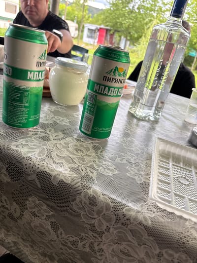 The image shows a table setting with several items on it. There are two cans of “PIRINSKO MLADO PIVO” beer, a bottle of Savoy vodka, a small jar filled with a white substance, and a white plastic container. The items are placed on a white lace tablecloth. Part of a person's face and hand are visible on the left, and another person wearing black is partially visible in the background. The table is outdoors, with trees visible in the background. The OCR on the cans indicates the product is beer and provides nutritional information in Bulgarian. The overall setting implies a casual gathering or picnic. The location, Dabovets, Bulgaria, is not evident from the image itself but inferred from the beer brand and OCR language.
