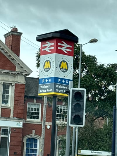 The image shows a signpost for Wallasey Grove Road station, a Merseyrail station in Wallasey, United Kingdom. The sign is red and white and features the Merseyrail logo. There is also a traffic light and a street lamp visible in the image. The station building is made of red brick and has several windows. Trees are visible in the background. The sky is overcast.
