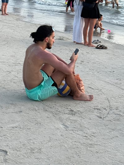 The image depicts a daytime scene on Cenang Beach, Malaysia. In the foreground, a male individual is seated directly on the light-colored sand, facing slightly right and away from the viewer. He possesses dark hair tied in a bun, a beard, and exhibits visible sunburn on his back and shoulders. He is shirtless and wears light blue swim shorts. A pair of sunglasses rests on his head. He holds a dark-colored smartphone in his right hand, oriented with the screen facing him. A blue and yellow volleyball is situated between his legs, and a textured, light brown item is partially discernible next to his right leg.

In the mid-ground and background, closer to the waterline, several other individuals are present. One person stands in white pants, while another person is seen in a black skirt and top, wearing a waist bag. A child is observed crouching near the water, holding a pink sand toy. Other indistinct figures are partially visible standing in the shallow ocean water or along the immediate shoreline. Two pairs of sandals, one black and white, and one brown, are resting on the sand near the water's edge. The environment consists of damp sand with minor ripples and footprints, leading to ocean water with small, gentle waves.