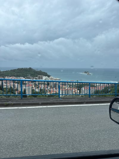 The image shows a view from a bridge in or near Stupovi, Montenegro. The Adriatic Sea is visible with a small rocky island. Coastal buildings with red-tiled roofs are interspersed with greenery on a hillside. The blue bridge railing runs along the side of the road, visible in the foreground. A car side mirror is present in the upper right corner. The sky is overcast with grey clouds.