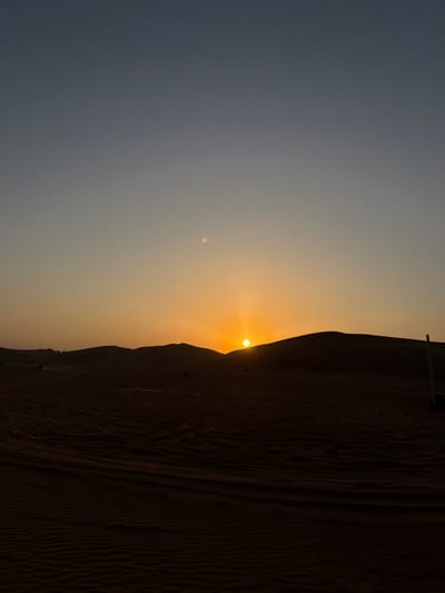 The image captures a desert sunset scene in Al Madam, United Arab Emirates. The sun, appearing as a brilliant orange disc, is partially visible above the horizon, cresting behind a dark sand dune in the right-center of the frame, emitting a strong orange glow across the lower sky. The sky above transitions from a clear blue-gray at the top to an orange-yellow hue near the horizon. A small, distinct bright circular object is present in the upper-middle left portion of the sky. The foreground and midground consist of dark, rippled desert sand, displaying visible tracks. Multiple undulating sand dunes form dark silhouettes against the bright horizon, extending across the entire width of the scene. In the far distance on the left, several faint, indistinct dark shapes are visible, potentially representing vehicles or people. On the far right side of the image, a light-colored vertical pole or object is partially visible, extending upwards from behind a dune. No specific human actions or interactions are discernible.