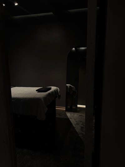 The image shows a dimly lit interior space in Ciudad López Mateos, Mexico. There is a cylindrical object to the left, possibly a pipe or post. Above, there is a reflective surface with an indistinct object on it. A box-shaped object with a patterned surface is visible in the foreground to the right, and it appears to have a dark opening on one side. The environment is dark, and the specific details are hard to distinguish.