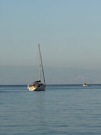 The image depicts a serene maritime scene. A white sailboat with its mast raised dominates the right side of the frame, floating on calm water. In the background, a distant boat is visible. The sky and the sea meet at the horizon, with a subtle coastline suggesting the location. The color palette is predominantly blue and white, reflecting the sky and water. There is no evidence to support the claim the image is in the city of Chianchitta, Italy.