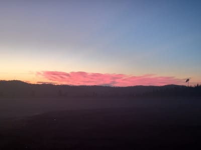 The image displays a twilight landscape in Santa Juana, Chile. The sky transitions from a light blue at the zenith to pale yellow and orange near the horizon. A prominent, elongated band of vibrant pink clouds stretches horizontally across the lower sky, positioned directly above a dark, undulating silhouette of hills densely covered with trees or foliage. The immediate foreground and midground are a dark, undifferentiated flat expanse, lacking specific detail due to the low light conditions. On the far right, a dark, pole-mounted object resembling a deflated windsock is visible against the sky, rising above the distant treeline. On the far left, a faint, slender vertical structure, possibly a utility pole or communication tower, is discernible above the contour of the hills. No individuals, animals, or specific activities are present.