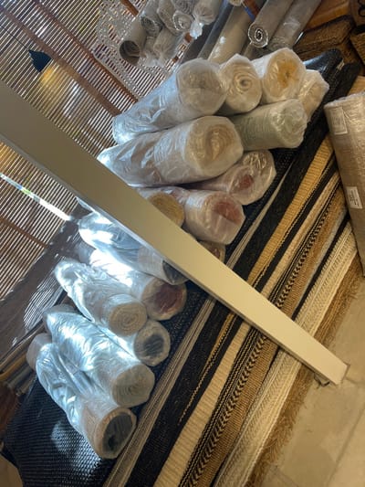 Rolled rugs wrapped in plastic are stacked, some organized on woven basket-like shelves and others resting on striped flat rugs. A long, white rectangular beam diagonally crosses the scene. Sunlight filters through a woven blind, casting shadows on some of the rolls. The rugs have varied colors and textures. Some have identifying labels attached to the packaging. The arrangement seems to be in a retail setting or a storage area.