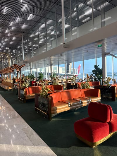 This image depicts a spacious airport terminal lounge or waiting area located in Roissy-en-France, France. The scene is illuminated by natural light entering through large, floor-to-ceiling windows on the right, which overlook an airport tarmac.
The environment features a high, multi-layered ceiling with a slatted, metallic design incorporating linear light fixtures. Below a structural beam, another ceiling section with recessed panel lighting is visible. A green exit sign with a right-pointing arrow is affixed above the windows. Small security cameras are mounted on ceiling elements.
The flooring transitions from polished white marble tiles with gray veining on the left to a dark green carpet covering the main seating area.
The primary furniture consists of multiple rows of interconnected, low-backed modular sofas and booths. These are predominantly two-toned, with the upper sections and back cushions upholstered in a burnt orange or reddish-orange fabric, while the lower seat cushions are in a tan or beige material. They feature brass-colored metal frames and armrests, some with circular black and white inserts, possibly power outlets or cup holders. Between some seating units, dark, possibly marble-clad planters with brass trim hold large green leafy plants, some displaying reddish undersides.
Further left, a distinctive "train car" or "booth" like structure with arched wooden elements and decorative hanging lights runs parallel to the main seating. One individual is partially visible standing near this structure.
In the immediate foreground on the right, a separate, single-seat modular lounge chair upholstered in a solid deep red velvet fabric with a brass-colored base is positioned on the green carpet.
Through the windows, a portion of an aircraft with a red tail and a jet bridge attached is visible on the tarmac under a clear sky, indicating active airport operations. The lounge area appears largely unoccupied.