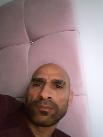 The image depicts a bald man with dark hair on his chin and a furrowed brow. He is lying on a pink, tufted headboard. A single button is visible on the headboard. The man is wearing a burgundy-colored shirt. The background consists of a plain white wall. The location is not discernible from the image.