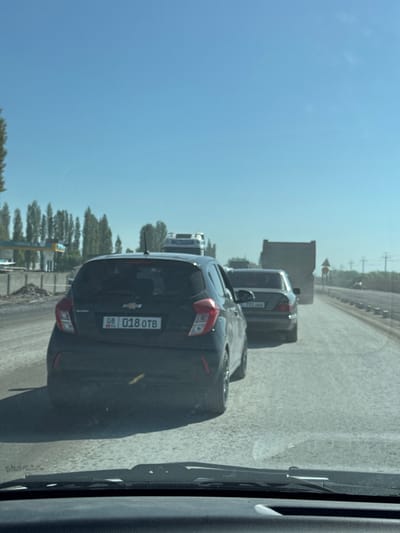 The image depicts a scene of road traffic, likely within or near the city of Tömönkü Norus, Kyrgyzstan, inferred from the license plate on the rear of a dark-colored Chevrolet vehicle showing "018 OTB" with the country code "KG" (Kyrgyzstan). The Chevrolet is positioned closely behind a grey vehicle, which is in turn behind a large truck. The view is from inside a vehicle, looking through a slightly dirty window. Outside the vehicle window, the road stretches ahead under a clear blue sky. Trees and some infrastructure are visible along the side of the road.