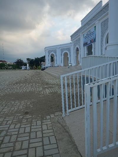 This image depicts the exterior of a large, modern white building, likely a mosque or community center, located in Meureudu, Indonesia, under an overcast sky.

The scene is dominated by the building, positioned to the right and center, featuring a multi-tiered entrance with numerous wide concrete steps leading to an elevated platform. The building's facade is white, adorned with intricate dark grey decorative patterns, notably around a series of large arched openings. A prominent green and white banner, displaying text in an Indonesian language and a group photograph, is affixed to the building's upper facade, suggesting an event. Near the base of the steps on the elevated platform, several illuminated stage lights or similar equipment are visible.

In the immediate foreground, a white metal railing curves into the right side of the frame, leading to a concrete ramp or pathway. The ground in front of the building is a paved area constructed from rectangular grey interlocking bricks, interspersed with patches of light brown dirt. To the left of the building, a few motorcycles or scooters are parked on this paved surface.

Further into the background on the left, an area of green foliage and trees is visible, with distant low-rise buildings featuring red roofs. A tall, slender communication or transmission tower extends vertically against the cloudy sky on the far left horizon. The sky itself is heavily overcast with dense grey cloud cover. No individuals are clearly discernible within the frame.