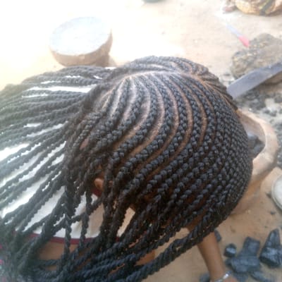 An overhead view captures the head of a person in Ibreda, Nigeria, featuring an elaborate hairstyle composed of intricate black cornrow braids that curve across the scalp, and numerous long, twisted hair extensions that hang downwards over the left shoulder. The person's left shoulder is covered by a white garment with red trim. A silver-colored bracelet is visible on their right wrist. The outdoor setting includes a light-colored ground surface, appearing to be dirt or concrete. In the background, objects on the ground include a circular flat stone, a dark wooden mortar or similar large object, several dark, irregularly shaped lumps (consistent with charcoal), a red-capped pen, and a dark elongated object.