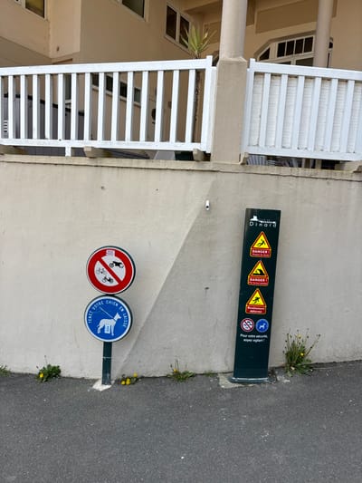 The scene is located in Dinard, France, at 48.636 latitude and -2.049 longitude.  A white fence runs along the top of a white concrete wall with an angled section.  The wall is in front of a building with a white wooden door. There are two signs on the wall. The left sign is blue and white and reads "Tenir votre chien en laisse" which means "Keep your dog on a leash." The right sign is red and white with black symbols and reads "Danger." The building is at the top of a hill and a sidewalk runs in front of it. The ground is a dark, gray asphalt. There are several plants and a few small flowers growing in the ground.  There are no people in the image.