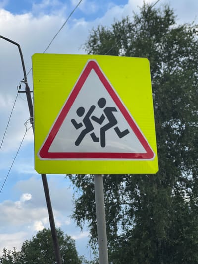 The image shows a triangular road sign with a yellow background and a red border. Inside the triangle, two black silhouettes of children are depicted running. The sign is mounted on a metal post. In the background, there is a pole with wires, lush green trees, and a sky with clouds. The scene appears to be an outdoor environment.
