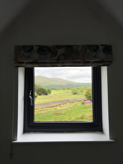 The image presents a view from inside a dimly lit room, looking out through a window onto a vibrant, natural landscape. The window itself has a dark, possibly black or dark grey, frame set within a light-colored, likely white, wall. To the left, a decorative Roman blind with a pattern of blue, green, and brown leaf-like shapes is partially pulled up. A silver-beaded pull chain for the blind is visible along the bottom edge of the window sill.

Outside the window, the scene depicts a lush, green, and hilly terrain under an overcast, cloudy sky, suggesting a typical day in Sconser, United Kingdom. Notably, the outdoor landscape is vertically oriented, appearing rotated 90 degrees clockwise. Rolling green hills dominate the background, while closer to the foreground are fields, scattered trees, and a distinct track or path running through the verdant expanse. A bright patch of purple or pink flowering bushes, possibly rhododendrons or heather, stands out on the right side of the outdoor view. There are no people visible, and no specific activity is taking place; it's a static view of the picturesque, rugged scenery. No visible text is present in the image.