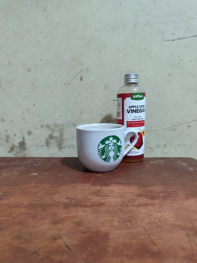 A white Starbucks mug, filled with a light-colored liquid, sits on a reddish-brown wooden surface. To the right of the mug is a bottle of Safiya Apple Cider Vinegar, with a red label. The background is a textured, off-white wall with some smudges and marks. The lighting appears to be artificial and casts a shadow from the mug and bottle onto the wooden surface. The scene is indoors, with no visible windows or outdoor elements. The overall impression is one of a still life composition, possibly staged in a home or small business environment within Lancokpanteara, Indonesia, though no specific city landmarks are visible.