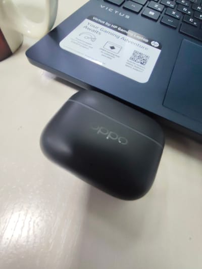 The scene depicts a desktop environment in Kangli, Uzbekistan. A dark gray HP Victus gaming laptop occupies the upper right portion of the frame, with its dark gray keyboard and a portion of its palm rest visible. A white rectangular sticker is affixed to the palm rest, displaying text that includes "Victus by HP Gaming Laptop" and "Your Gaming Adventure Awaits," alongside an HP logo and a QR code. Directly in front of the laptop, a dark gray OPPO brand wireless earbud charging case rests on the surface; the "oppo" logo is subtly embossed on its lid. In the upper left corner, a partial view of a white ceramic mug with vertical red or dark stripes is visible. All objects are stationary and positioned on a light-colored, possibly white or off-white, textured surface, likely a desk or table. No individuals are present in the image.